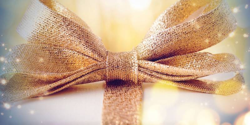 A golden bow sitting on top of a white present with the golden ribbon wrapped around it is surrounded by sparkles.